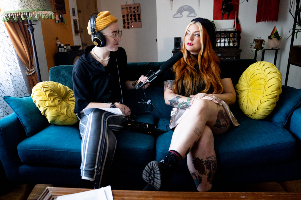 Ro Kelly, a white woman weawring a yellow beanie and black shirt with striped pants interviews Auralie Wilde, a white woman with a black beret, shirt, and pink skirt on.