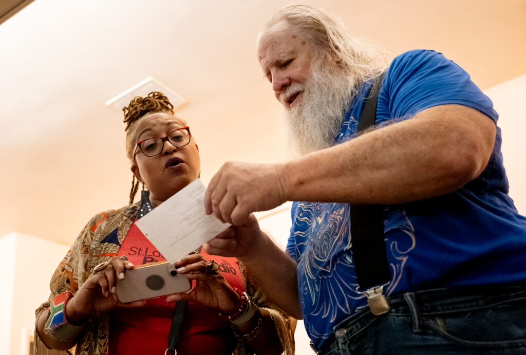 A Black woman leans over to look at a call card being held by a white man in a blue shirt and black suspenders.