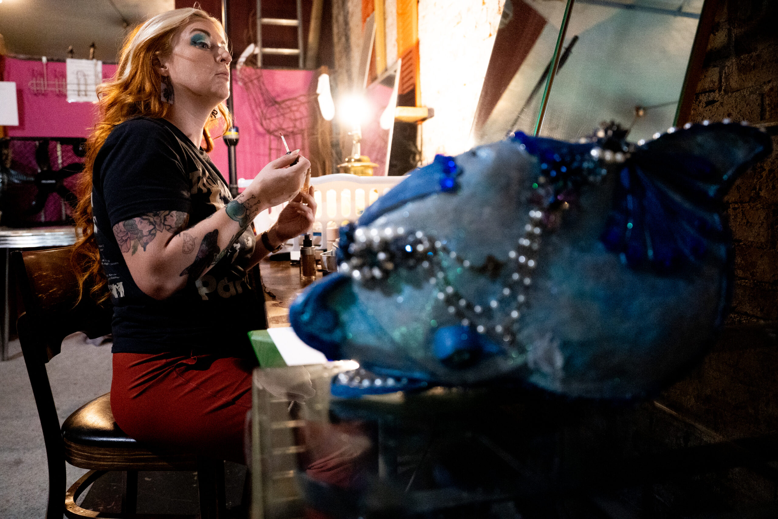 A white woman looks in the mirror putting on make-up in a dimly lit, makeshift dressing room. A closeup of a dolphin head is in the foreground.