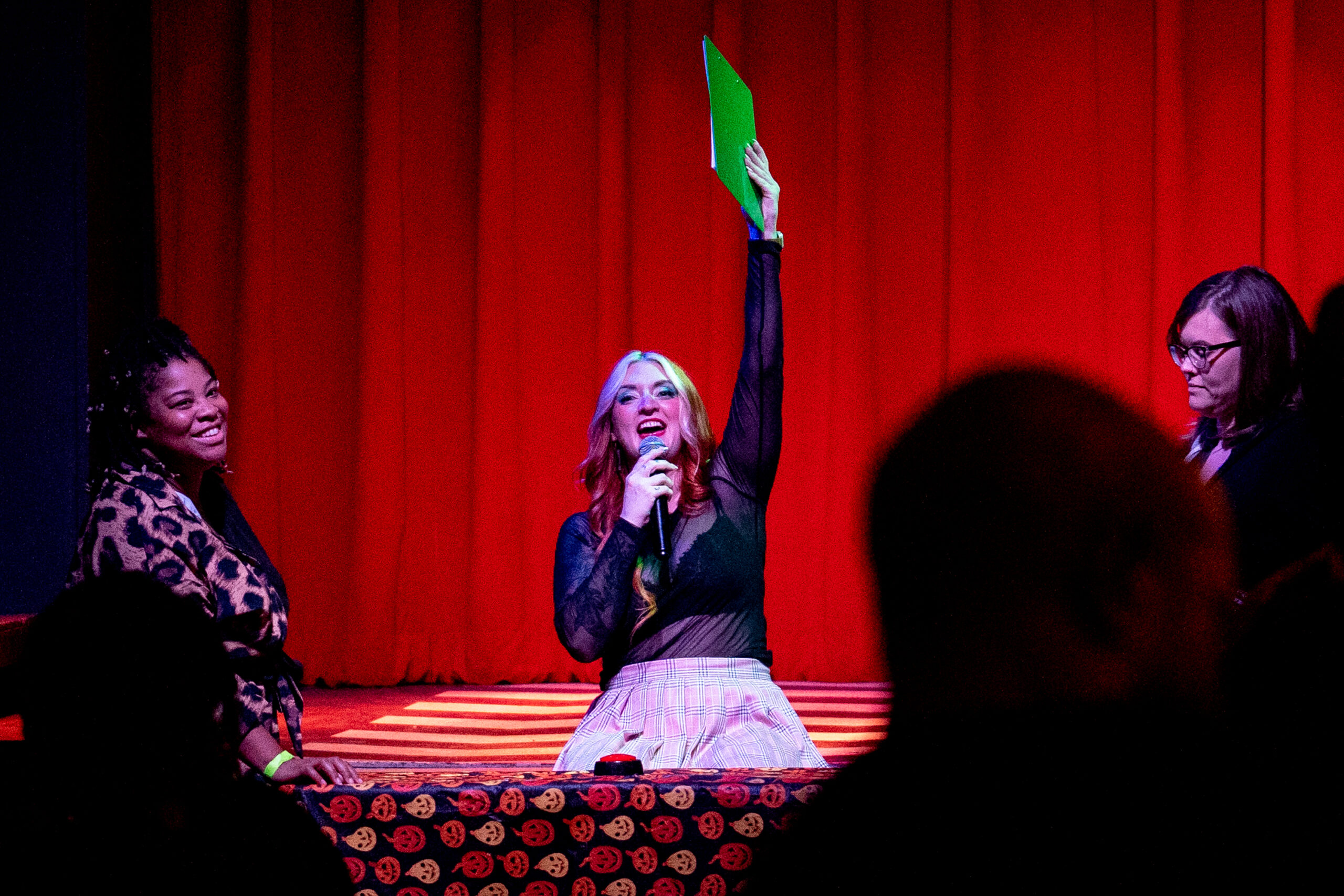 A white woman sitting in front of a table raises a green clipboard above her head in front of a stage with two smiling game participants on each side of the table.