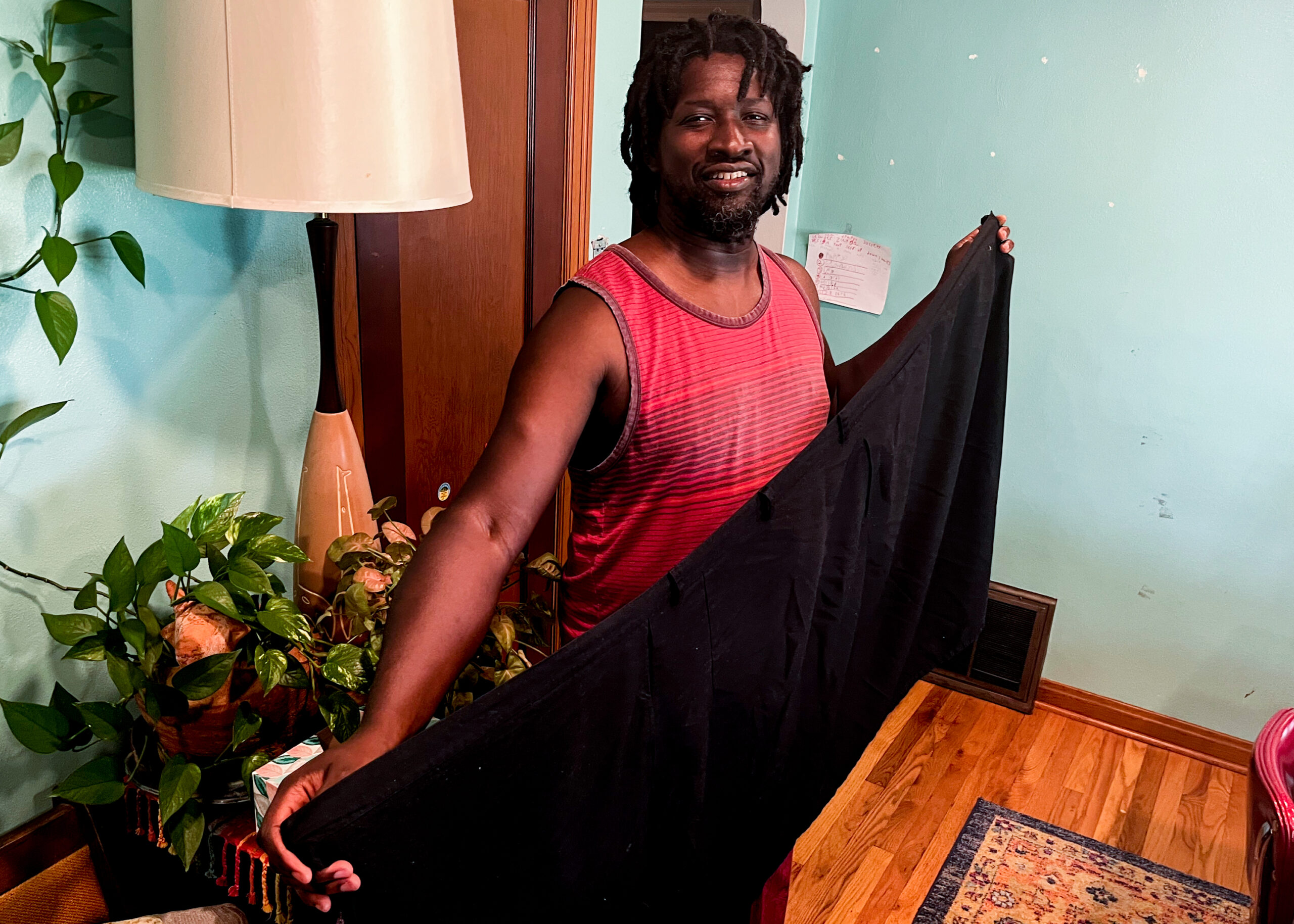 A Black man in a red shirt holds up a black wrap skirt.