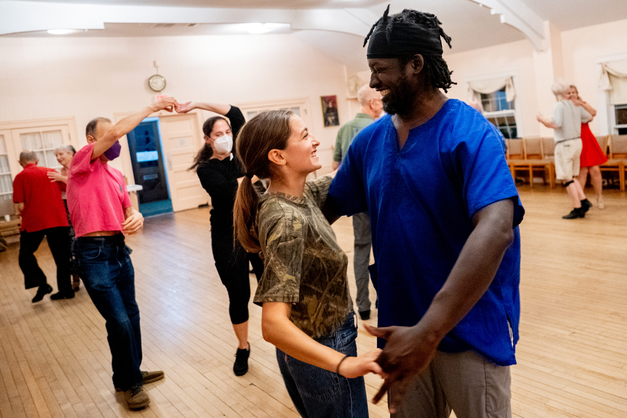 A Black man in his early 40s in a blue shirt dances with a white woman in her 20s wearing a camouflage shirt.