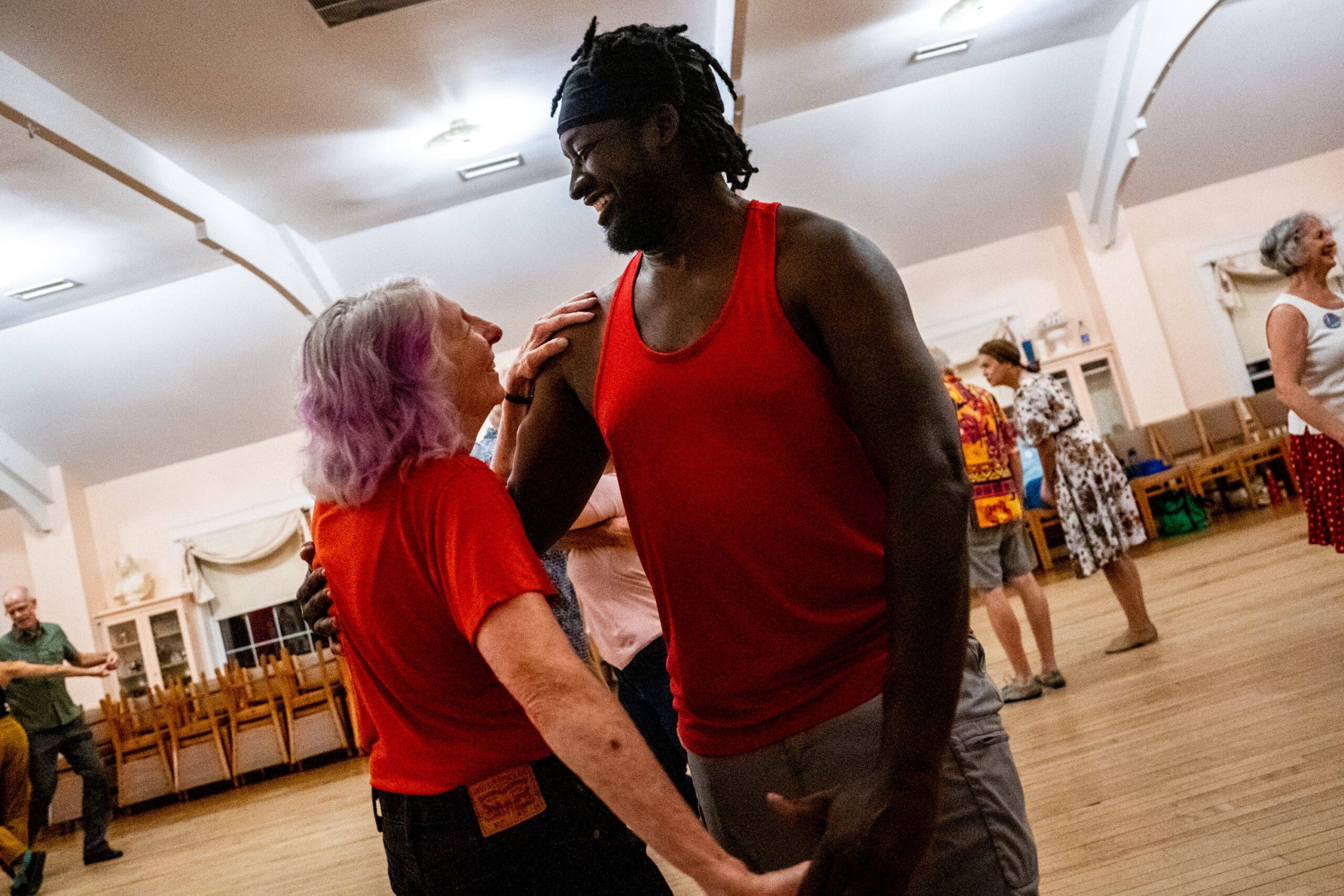 A Black man in his early 40s in a red shirt dances with a white woman in her mid-70s wearing a red shirt.
