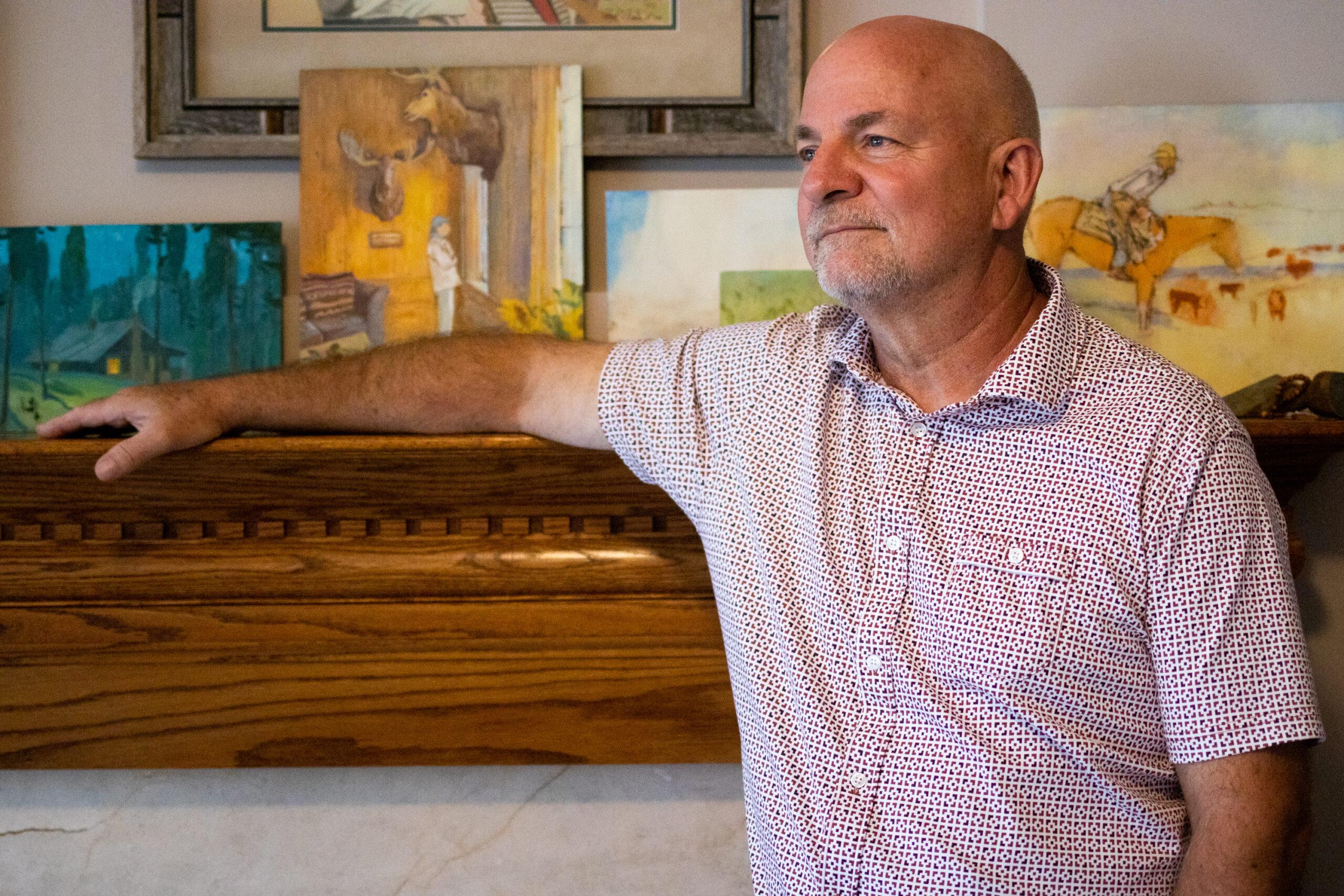 A white man in his 60s stands casually with one arm draped over the top of a fireplace mantle. On the mantle, a number of his oil paintings depicting scenes of the American West are informally displayed.
