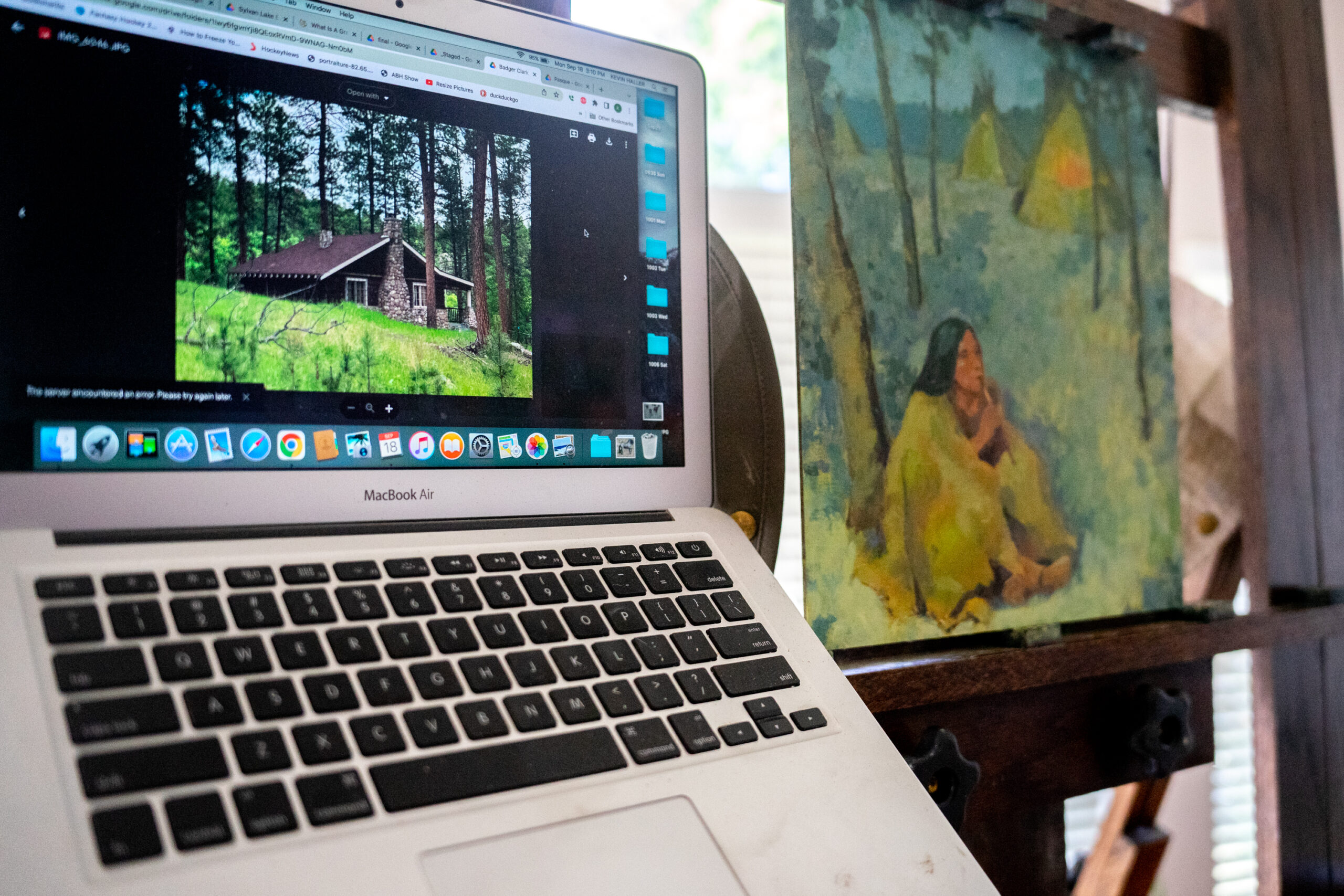 A computer with an image of a log cabin sits beside an in-progress painting of teepees on a wooden easel in an art studio.