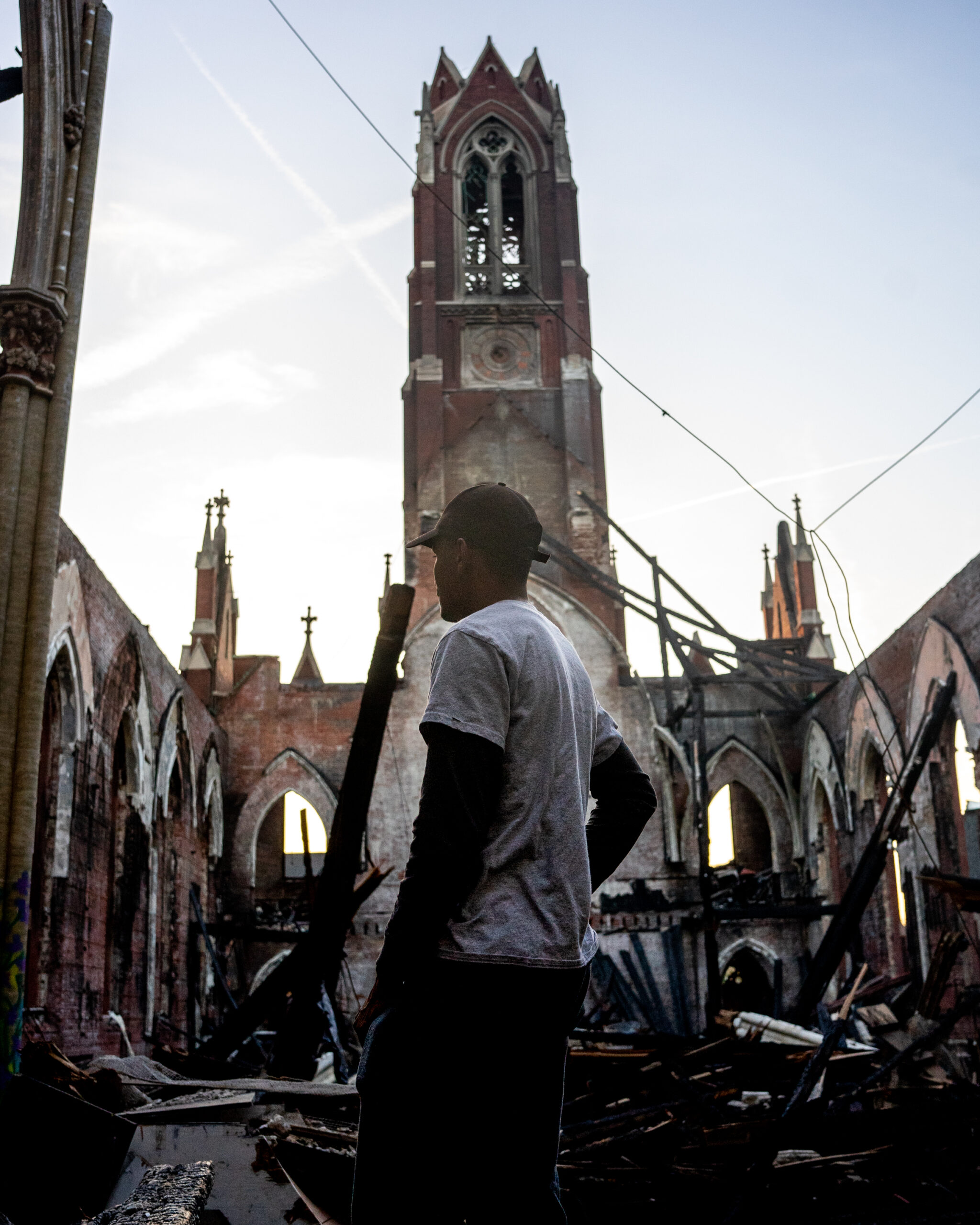 A young Black man in a baseball cap stands inside the remains of a church sanctuary, silhouetted against the steeple and the sky.