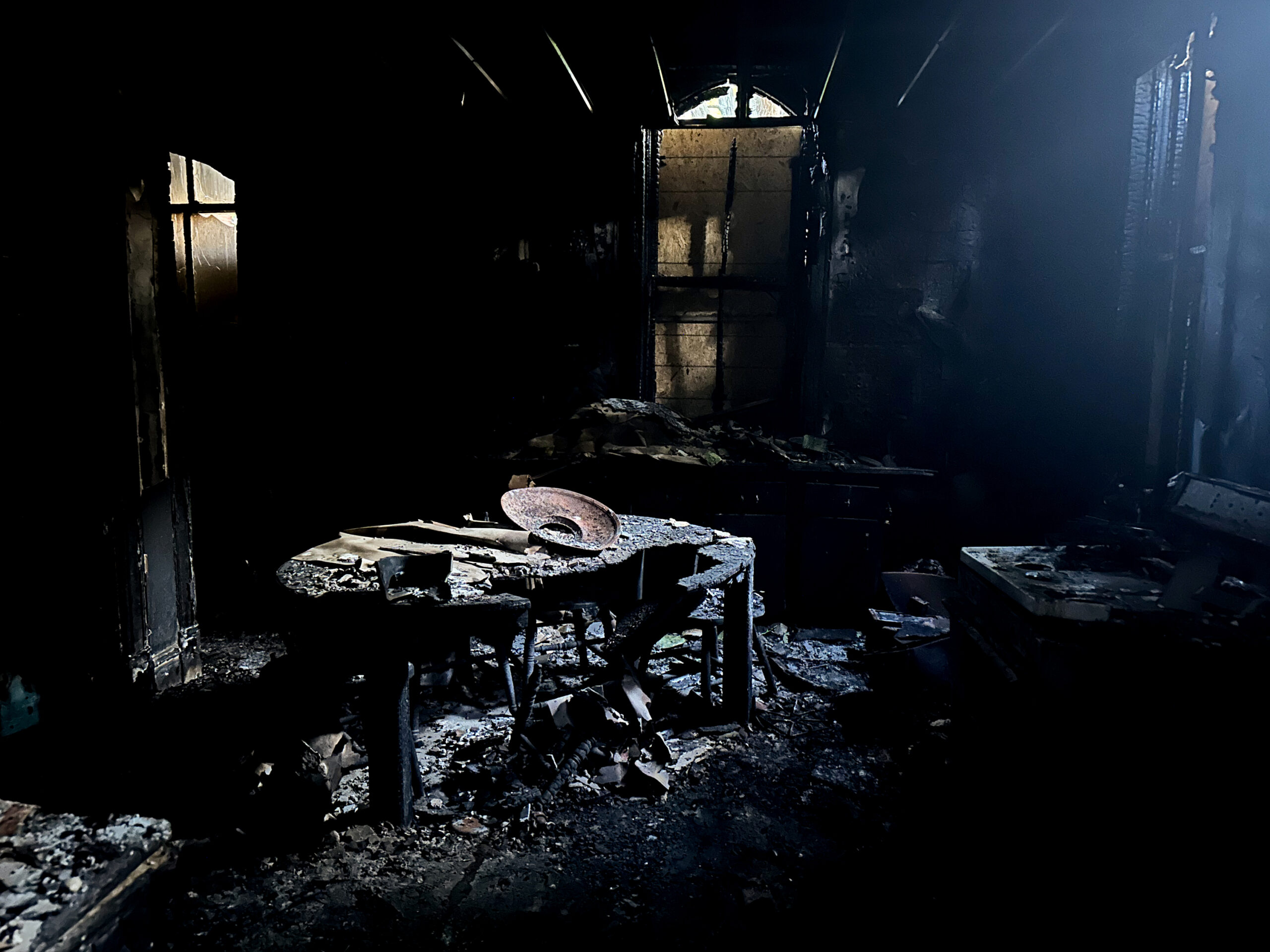 Light falls from a broken window onto the charred remnants of an oven and a kitchen table.