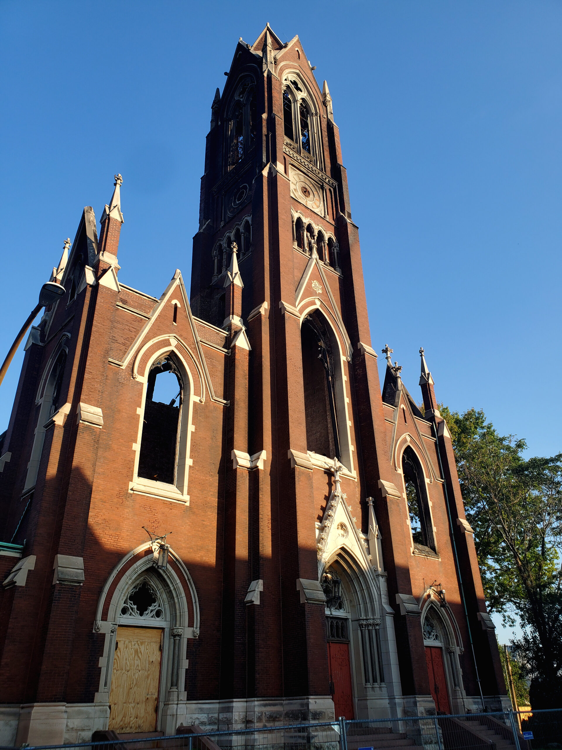 A red brick church with a boarded up door, scorch marks, and broken windows is visible in front of a blue sky.