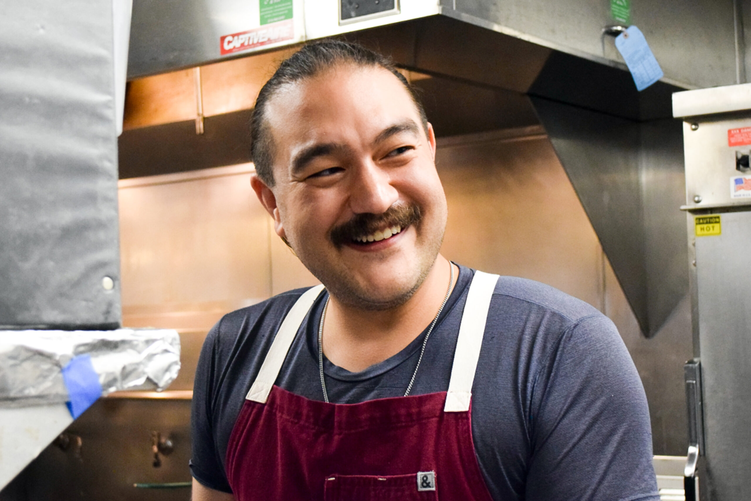 A close up of a middle aged fair-skinned man in a commercial kitchen smiling at someone off camera.